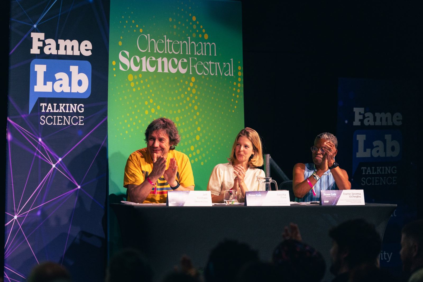 Three judges applauding at a table on stage, in front of FameLab and Cheltenham Science Festival banners