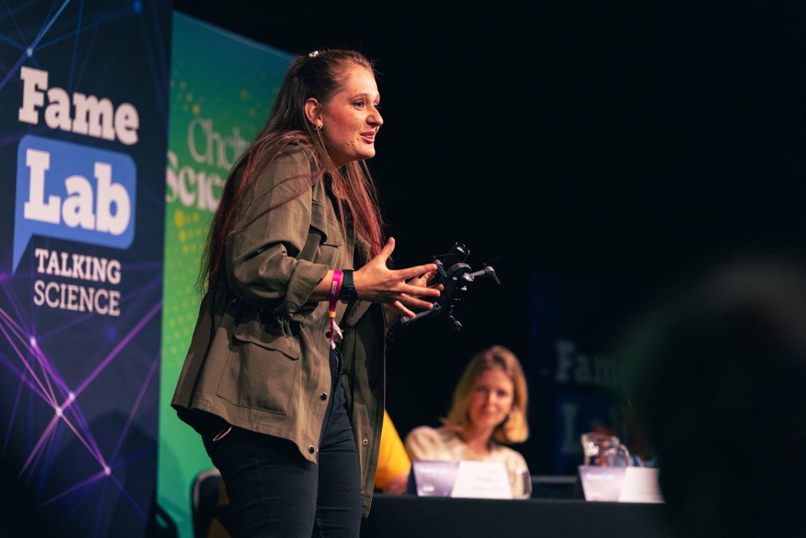 A FameLab finalist on stage holding a drone as a prop, mid-talk, with the judges' table behind her
